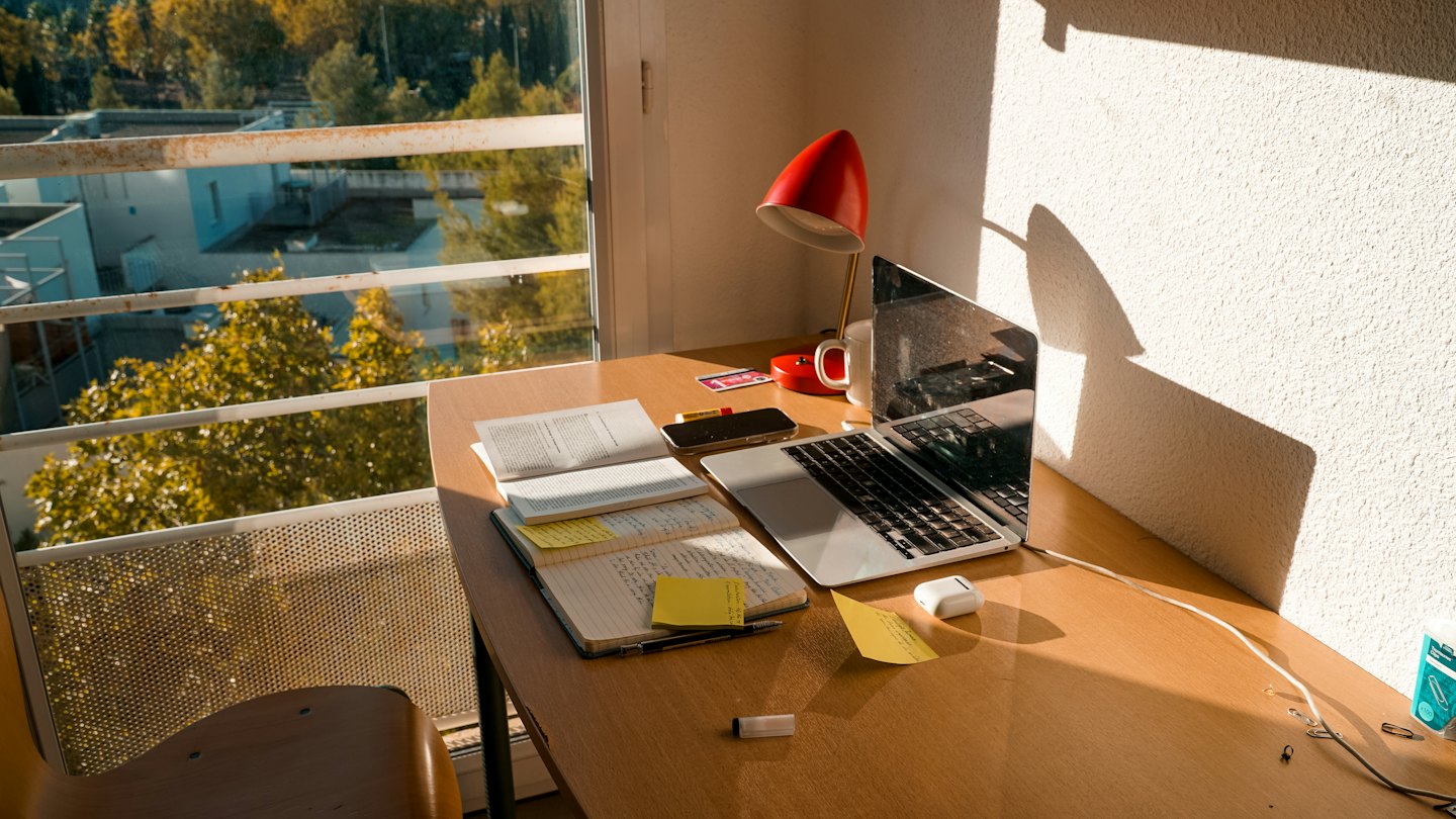 Open laptop and books on a wooden desk near a window.