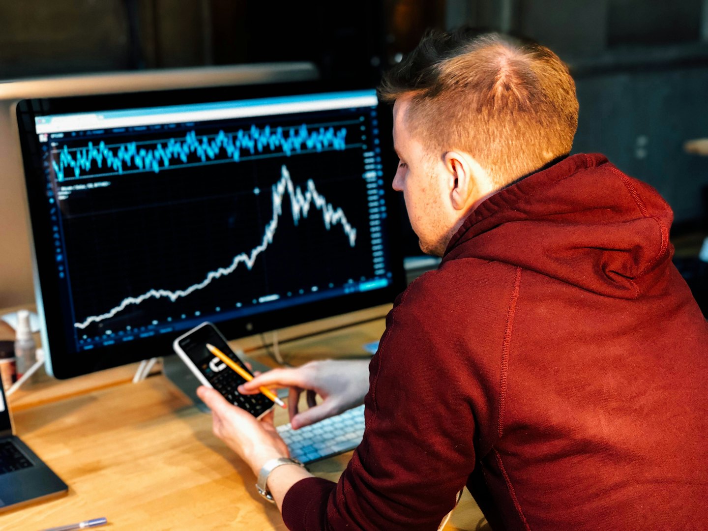 Man in hoodie studies financial charts on computer and phone.
